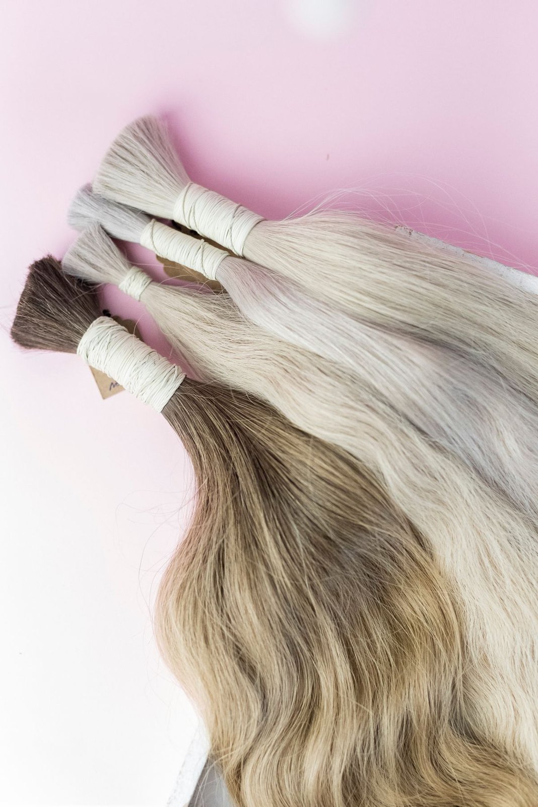 Losse blonde en bruinblonde haarbundels met witte binddraad op een zachtroze ondergrond.